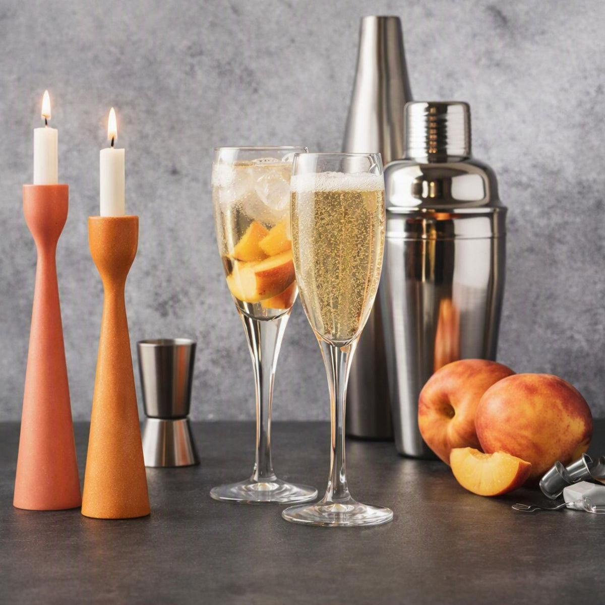 Two lit tapered candle holders in terracotta and orange stand beside two filled Prosecco flutes with peach slices, a metal jigger, and a stainless steel cocktail shaker on a dark tabletop against a gray textured backdrop. Gives the feel for the fragrance oil Prosecco and Peaches.