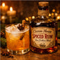 A bottle of Cocoon House Spiced Rum next to a glass of the amber-colored apple spiced cider cocktail, garnished with a cinnamon stick and star anise, set on a rustic wooden table with warm blurred candles in the background to represent Apple Cider Spiced Rum Fragrance Oil.