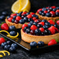 Three berry tartlets topped with fresh strawberries and blueberries on a black rectangular plate, with lemon slices in the background on a dark surface. Used to give the idea of Berrylicious Fragrance Oil.