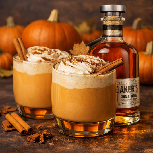 Two glasses of an Bourbon Ppumpkin spice latte topped with whipped cream and cinnamon sticks, styled with a bottle of Caker's Single Barrel bourbon and pumpkins in the background to represent Bourbon Pumpkin