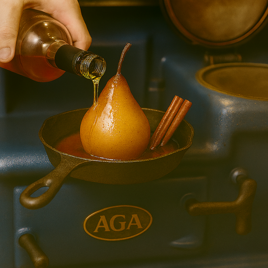 Poached pear in cast iron skillet with cinnamon sticks, brandy being poured over top, set on a deep blue AGA stove. Rich amber sauce pools around the pear, highlighting its glossy caramelized surface.