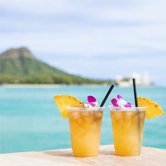 Two clear cups filled with an iced golden drink, garnished with pineapple slices and purple orchids, set on a wooden table with a blurred tropical beach and mountain background for Butt Naked Fragrance Oil.