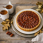 A large pecan pie on a white plate with a rustic metal charger plate, styled on a wooden table with two white mugs of coffee, scattered autumn leaves, and loose pecans. Smells like Butter Pecan Pie Fragrance Oil from Northstar.