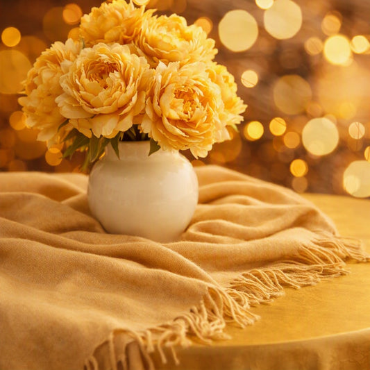 A vase of golden peonies in a round white vase on top of a gold cashmere shawl with round golden lights used to represent Cashmere Glow Fragrance Oil.