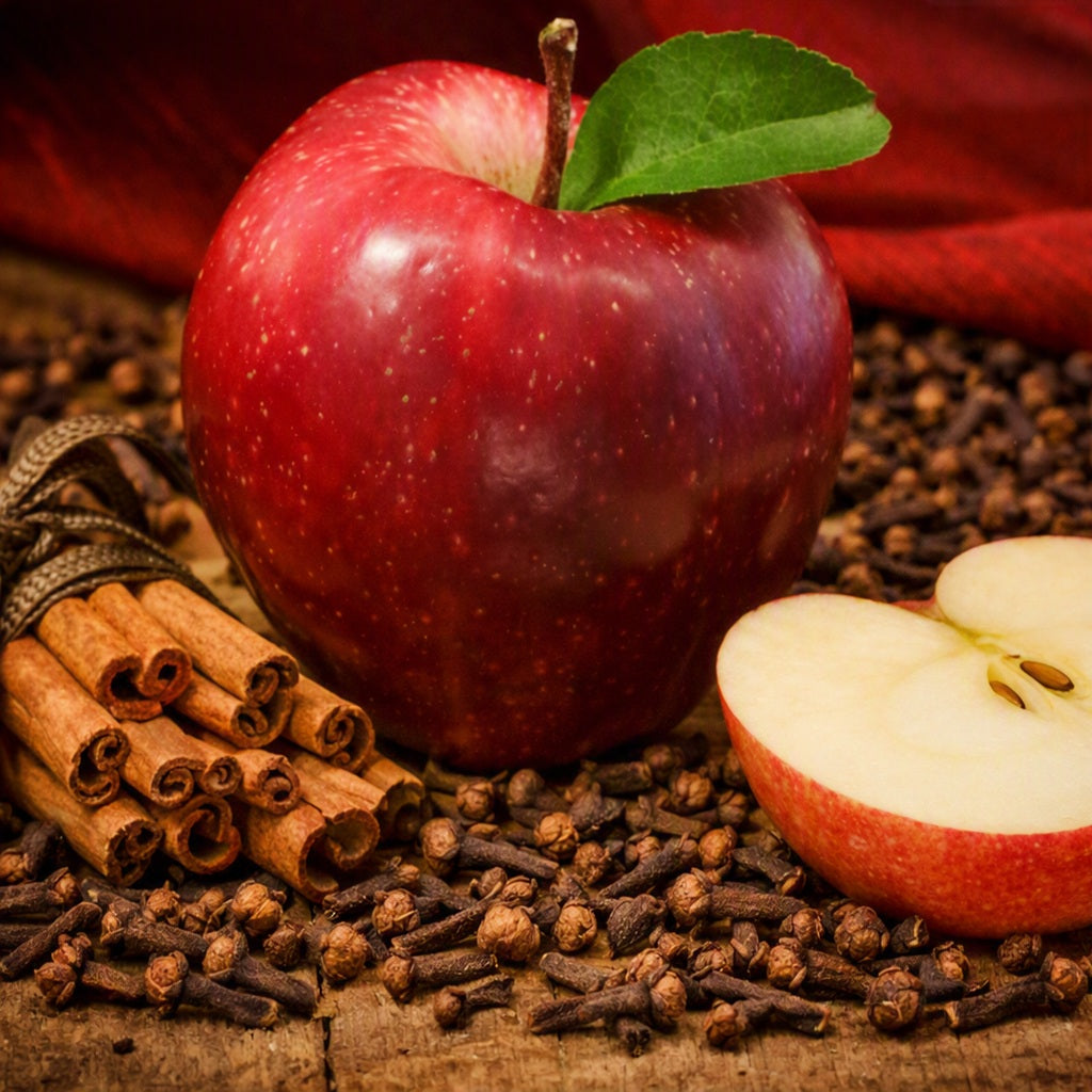 A glossy red apple with a green leaf sits on a rustic wooden surface surrounded by cinnamon sticks, whole cloves, and a sliced apple half, with a dark red fabric backdrop. Gives the idea of the fragrance oil Cinnamon N Spiced Apple.