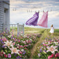 A woman in a white dress stands on a grassy path between flower beds, looking toward two flowing dresses hanging on a clothesline outside a white house. The scene is filled with pink, purple, and white flowers under a cloudy sky. Represent the fragrance oil Clothesline Fresh.