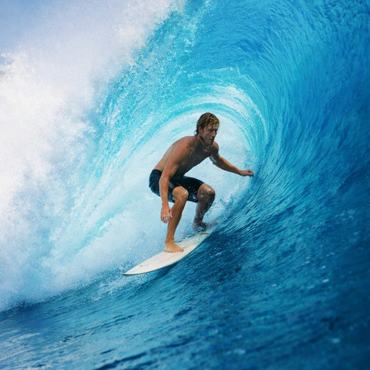 Cool Water Fragrance Oil is represented by A shirtless male surfer crouches on a white surfboard inside a large curling blue ocean wave, with white sea foam visible on the left.