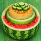 A decorative fruit bowl carved from a watermelon half, holding a sliced cantaloupe and topped with cucumber rounds, set on a green wooden surface to signify Cucumber Melon Northstar Fragrance Oil