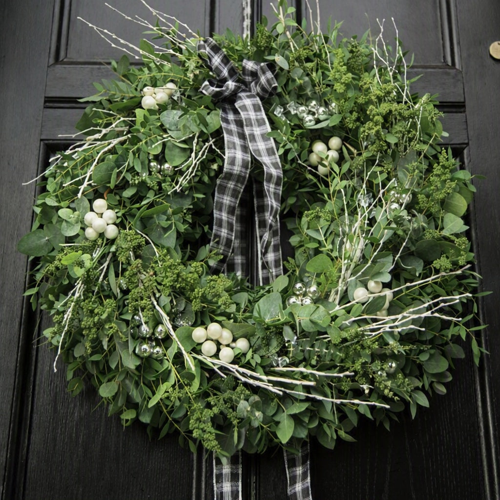 A green holiday wreath with eucalyptus leaves, white berries, and white twig branches, tied with a black and white plaid ribbon bow, hanging on a black paneled door to give the idea of Dried Eucalyptus Fragrance Oil.