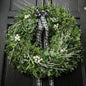 A green holiday wreath with eucalyptus leaves, white berries, and white twig branches, tied with a black and white plaid ribbon bow, hanging on a black paneled door to give the idea of Dried Eucalyptus Fragrance Oil.