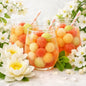 Three clear glasses filled with a colorful melon drink and pink striped straws, surrounded by white flowers and mint leaves on a light surface to look like Fresca Melonade fragrance oil smells.