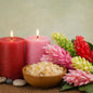 Two lit pink and red candles beside a wooden bowl filled with bath salts, surrounded by pink and red flowers on a burlap surface against a soft beige background. To represent the smell of Ginger Frankincense Fragrance Oil.