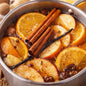 A close-up of a stainless steel pot filled with a warm spiced drink or syrup mixture, featuring sliced oranges, cinnamon sticks, vanilla beans, and apple slices in a glossy brown liquid. This smells like Harvest Spice Fragrance Oil.