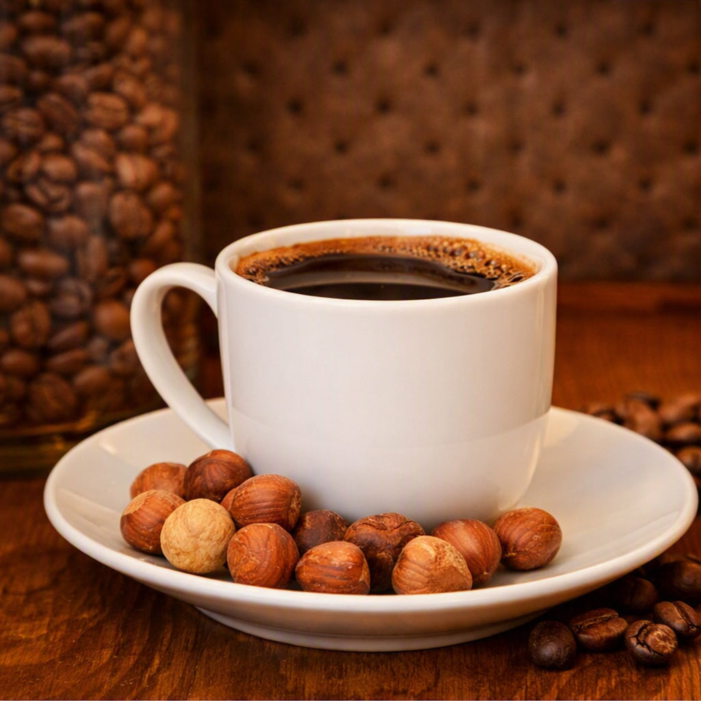 A white ceramic coffee cup filled with black coffee sits on a matching saucer, surrounded by whole hazelnuts and scattered coffee beans on a wooden surface, with a blurred background of more coffee beans and a brown tufted backdrop. Smells like the fragrance oil Hazelnut Coffee.