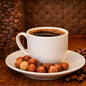 A white ceramic coffee cup filled with black coffee sits on a matching saucer, surrounded by whole hazelnuts and scattered coffee beans on a wooden surface, with a blurred background of more coffee beans and a brown tufted backdrop. Smells like the fragrance oil Hazelnut Coffee.