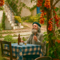 Elderly man with white beard and green cap sitting at a table with a blue and white checkered tablecloth in a rustic courtyard, surrounded by hanging tomato fruits, stone steps, and lush greenery, evoking a serene Mediterranean garden atmosphere and smelling a tomato leaf between his fingers.  Just like the Heritage Tomato Leaf fragrance oil.
