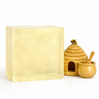   
A large translucent honey melt and pour soap block displayed beside a small woven beehive, a ceramic honey pot with a honey dipper, and a decorative bee perched on the hive. The soap has a warm golden color with smooth, glossy surfaces, and the scene is arranged on a bright white background to emphasize the honey theme and natural ingredients.