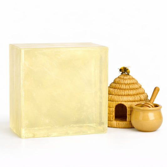   
A large translucent honey melt and pour soap block displayed beside a small woven beehive, a ceramic honey pot with a honey dipper, and a decorative bee perched on the hive. The soap has a warm golden color with smooth, glossy surfaces, and the scene is arranged on a bright white background to emphasize the honey theme and natural ingredients.
