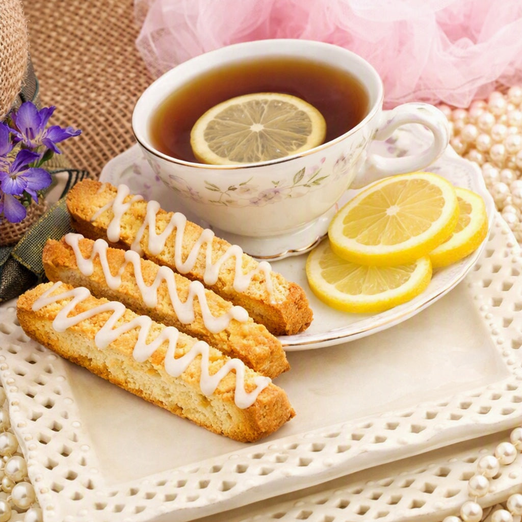 Three golden-brown Iced Lemon biscotti cookies with white icing drizzle are arranged on a white plate next to a white teacup filled with tea and a lemon slice, with lemon slices and a pink tulle cloth in the background. What Iced Lemon Biscotti Fragrance Oil smells like.