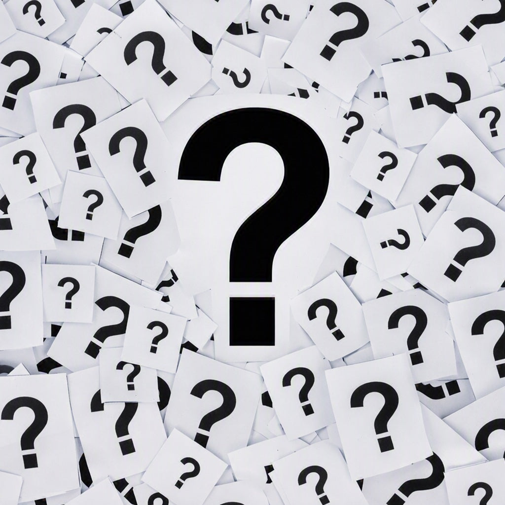 A central large black question mark on a white card, surrounded by many scattered white cards, each printed with a black question mark. Gives the idea of It's A Mystery Fragrance Oil scent.