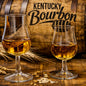 Two clear whiskey glasses filled with amber bourbon placed on a wooden surface in front of a wooden barrel printed with the words Kentucky Bourbon. Wheat stalks and a few grains are scattered around the glasses. This feels like the smell of Kentucky Bourbon Fragrance Oil.