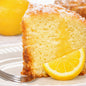 A close-up of a slice of lemon cake on a white plate with a fork, garnished with a lemon wedge, with a whole lemon blurred in the background. Gives the feel of Lemon Pound Cake Fragrance Oil.
