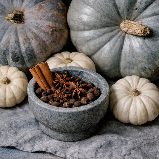 A gray stone mortar and pestle filled with whole spices, including cinnamon sticks and star anise, styled with small white pumpkins and larger blue-gray pumpkins on a textured gray fabric background. Gives the feel of Mahogany Pumpkin Fragrance Oil