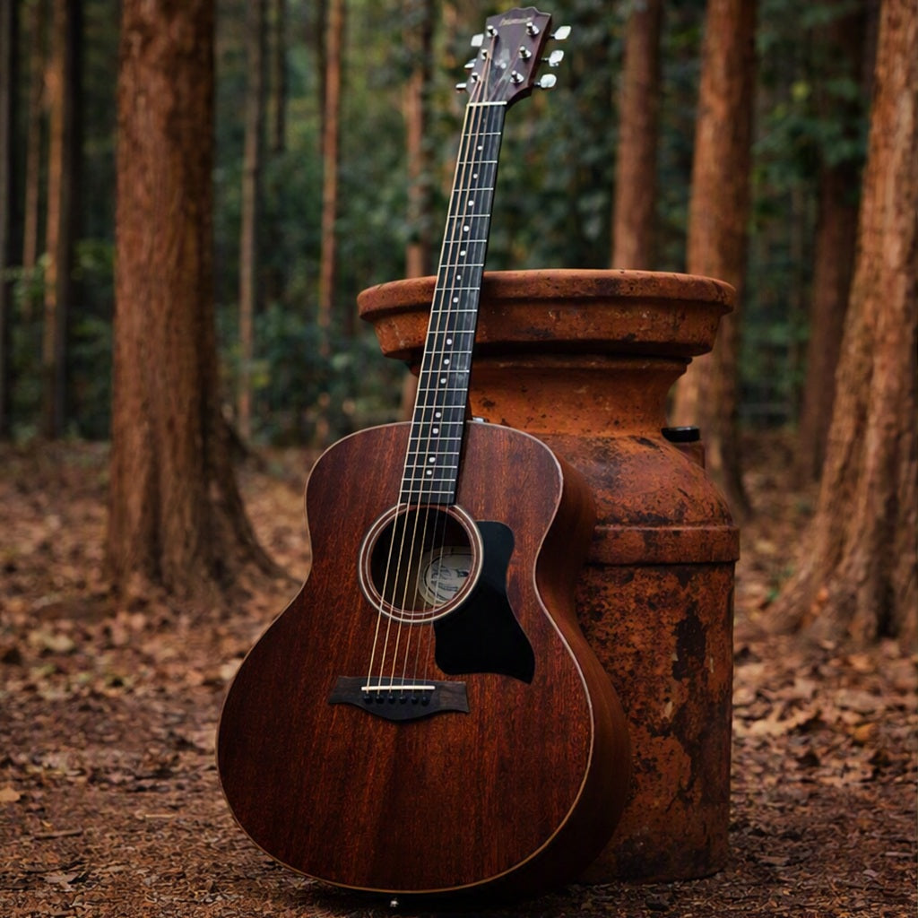 A dark brown mahogany acoustic guitar with a black pick guard leans against a large rusted metal milk can in a teak forest setting with trees and leaf-covered ground. Represents Mahogany Teakwood Fragrance Oil.