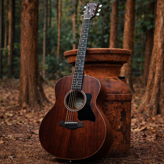 A dark brown mahogany acoustic guitar with a black pick guard leans against a large rusted metal milk can in a teak forest setting with trees and leaf-covered ground. Represents Mahogany Teakwood Fragrance Oil.
