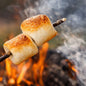 Two toasted marshmallows on a wooden skewer, roasting over an outdoor campfire with visible flames and smoke in the background. Gives the idea of Marshmallow Fireside Fragrance Oil.