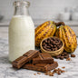 A glass milk bottle next to toasted cocoa beans and pods with milk chocolate pieces stacked in front.  Meant to give the idea of Milk Chocolate Fragrance Oil.
