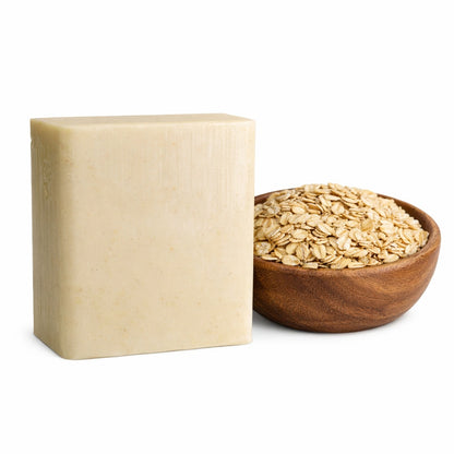 A creamy beige rectangular oatmeal soap bar sits beside a small wooden bowl filled with rolled oats, both placed on a clean white background. The soap has a smooth surface with subtle speckles, while the bowl of oats adds a natural, textured contrast.