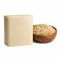 A creamy beige rectangular oatmeal soap bar sits beside a small wooden bowl filled with rolled oats, both placed on a clean white background. The soap has a smooth surface with subtle speckles, while the bowl of oats adds a natural, textured contrast.