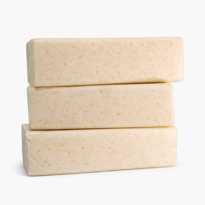 Three creamy oatmeal melt and pour soap base bars stacked horizontally against a clean white background. Each bar has smooth surfaces with rounded edges and visible golden specks throughout, highlighting the natural oatmeal texture and handcrafted quality.
