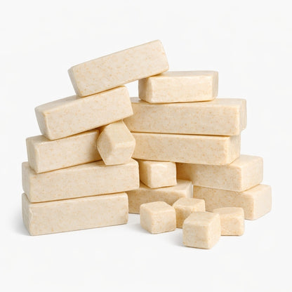 A loose stack of thirteen creamy oatmeal melt and pour soap base bars with a few small cubes scattered nearby, all set against a clean white background. The bars and cubes have smooth surfaces with visible golden specks, highlighting their natural oatmeal texture.