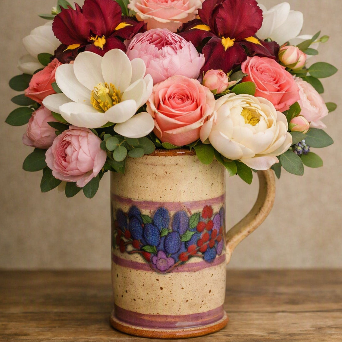 A ceramic mug filled with a large bouquet of pink, white, and burgundy flowers, including roses, peonies, and anemones, set on a wooden surface against a neutral beige background. To give the feel for the fragrance oil Perfect Petals