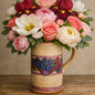A ceramic mug filled with a large bouquet of pink, white, and burgundy flowers, including roses, peonies, and anemones, set on a wooden surface against a neutral beige background. To give the feel for the fragrance oil Perfect Petals