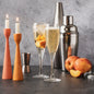 Two lit tapered candle holders in terracotta and orange stand beside two filled Prosecco flutes with peach slices, a metal jigger, and a stainless steel cocktail shaker on a dark tabletop against a gray textured backdrop. Gives the feel for the fragrance oil Prosecco and Peaches.