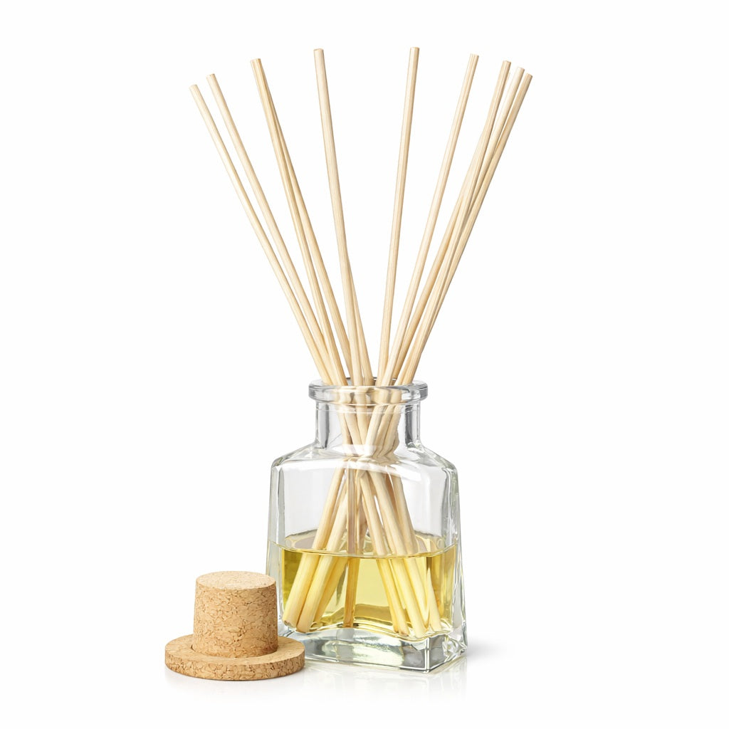 Clear glass reed diffuser bottle with a triangular base, filled with pale yellow fragrance oil and diffuser base. Several natural beige reeds fan out from the neck, and a cork stopper rests on a small cork disc beside the bottle. The setup is centered against a pure white background with soft, even lighting.