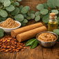 Natural cosmetic or skincare ingredients arranged on a wooden surface, including a bowl of light brown powder, a bowl of thick beige cream, two wooden sticks, scattered bark pieces, a small glass bottle of golden oil, and green eucalyptus leaves. What Sandalwood Eucalyptus fragrance oil smells like