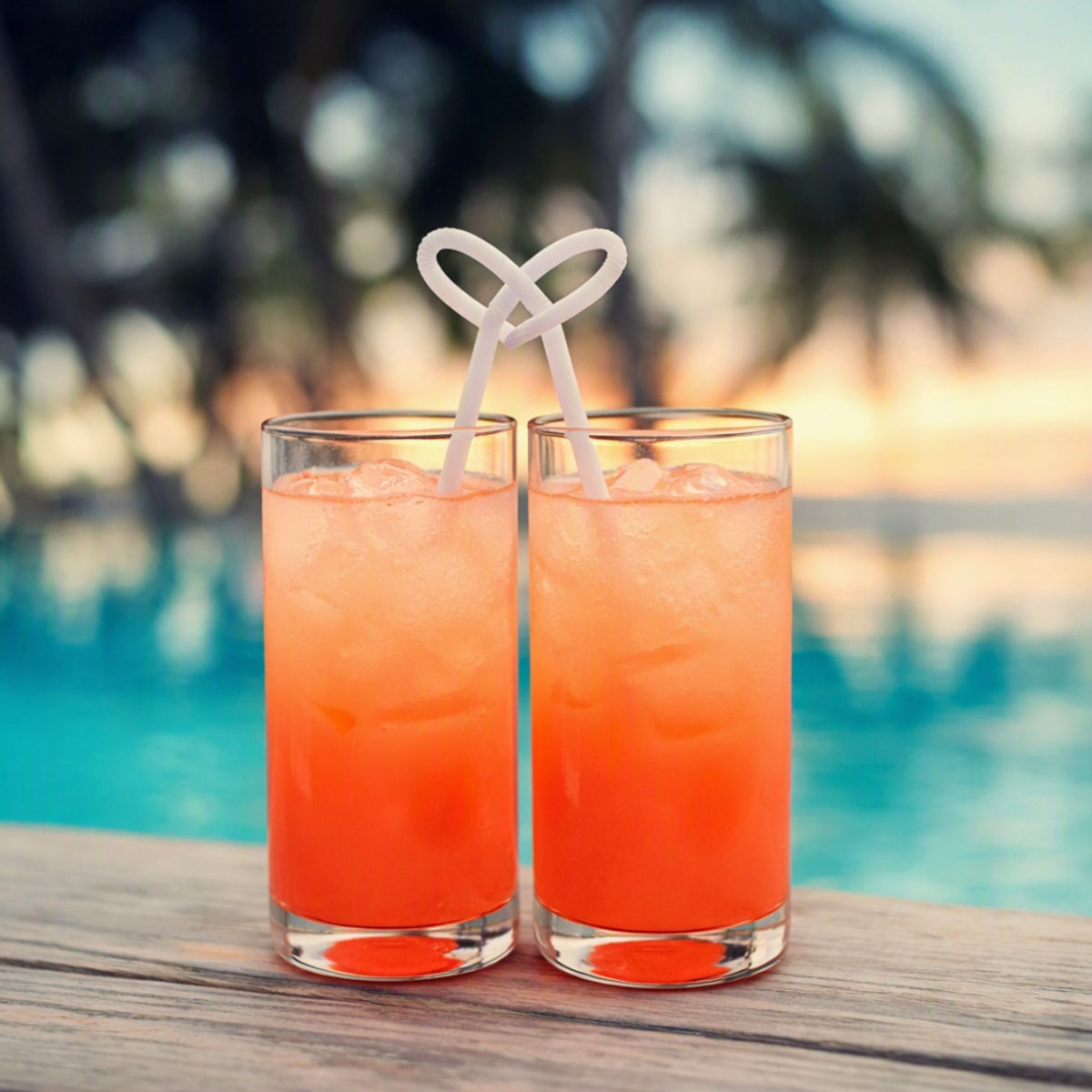Two tall clear glasses filled with an orange-red iced drink, each with a white looped straw, set on a wooden surface with a blurred poolside background. Made to feel like Sex on the Beach Fragrance Oil.