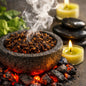 A stone mortar filled with whole cloves and tobacco, set over glowing coals with rising smoke, surrounded by candles, stones, and green leaves in a spa-like arrangement. Made to feel like Smoked Clove and Tobacco Fragrance Oil Smells.