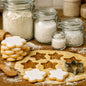 Star-shaped sugar cookies with white icing and metal star cookie cutters on a wooden surface, with glass jars of flour and sugar in the background. To give the feel for the smell of Sugar Cookie Fragrance Oil.