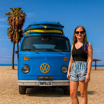 Young woman in black tank top and blue shorts smiling beside a bright blue vintage VW van with surfboards on the roof, parked on a sandy beach under a clear sky — inspired by Surf’s Up™ Fragrance Oil.