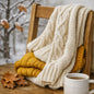 A cozy knit throw blanket in cream is draped over a wooden chair next to a folded mustard yellow knit blanket, with a white mug on a wooden table and a snowy outdoor scene blurred in the background. If Sweater Weather had a scent it would be this Fragrance Oil.