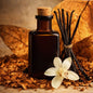 An amber glass bottle with a cork stopper stands on a bed of wood chips, surrounded by dried brown leaves, vanilla beans, and a white orchid flower. Used to give the feel for Tobacco Vanilla Fragrance Oil.