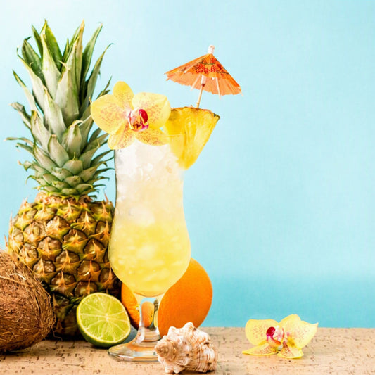 A tropical pineapple cocktail in a tall glass, garnished with a yellow orchid flower and a small paper umbrella, styled with a whole pineapple, lime, orange, coconut, and seashells on a light blue background. This is what Tropical Island Colada fragrance oil from Candle Cocoon smells like.