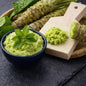 Fresh green wasabi mash in a dark blue bowl with mint leaves, alongside a wooden cutting board holding a piece of wasabi and a scoop of mashed wasabi, set on a dark slate surface. Gives the feel for Wasabi Mint Fragrance Oil by Northstar.