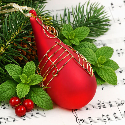 A glossy red Christmas ornament shaped like a teardrop with gold wire musical note patterns, decorated with a gold ribbon loop, styled with green pine branches, mint leaves, and red berries on sheet music. Smells like Winter Mint Fragrance Oil from Northstar.