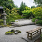 A serene Japanese-style garden with white raked gravel, a stone lantern, a wooden bench, mossy rocks, and dense green trees in the background. Gives the idea for the smell of Zen Garden Fragrance Oil